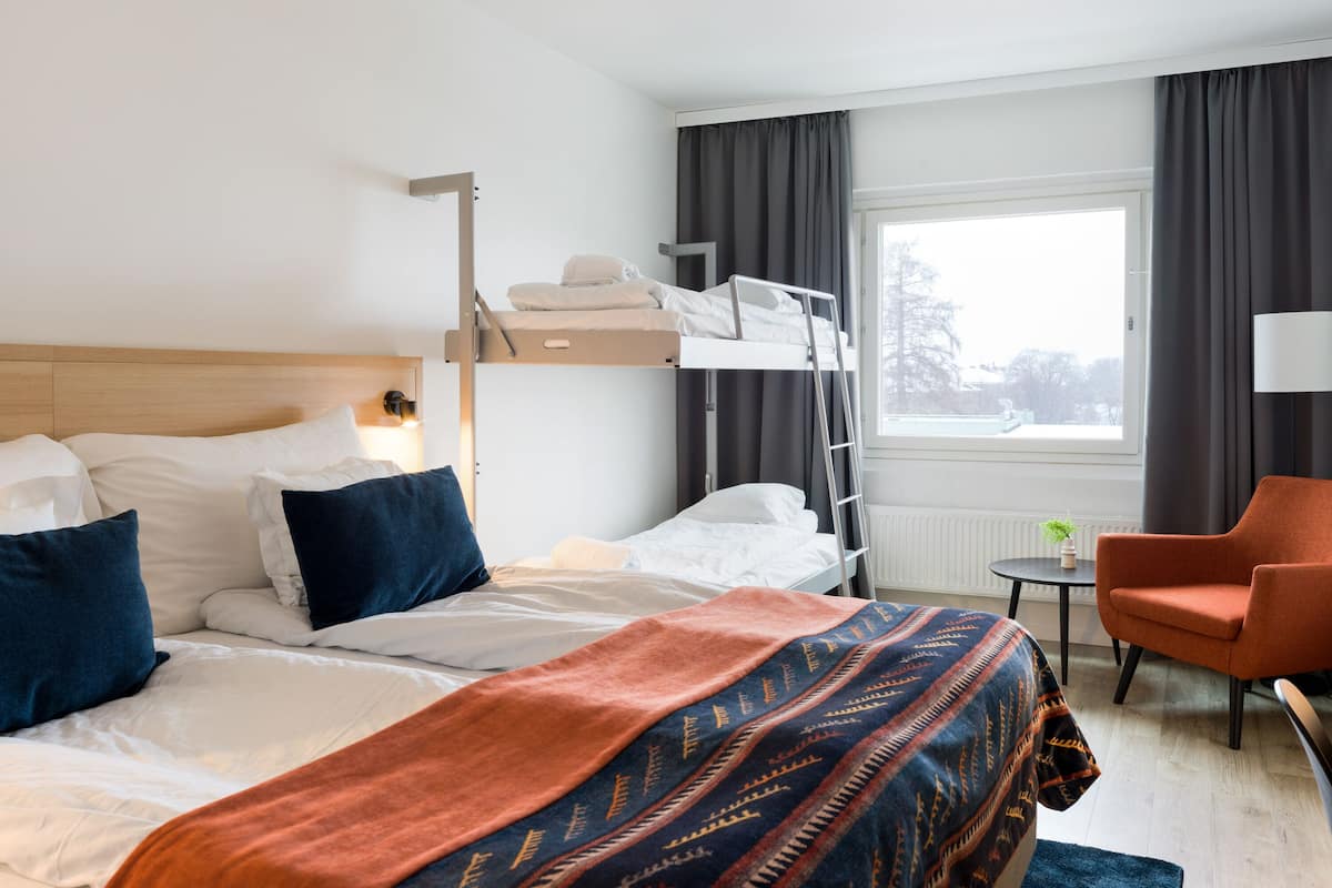 Hotellihuoneessa on kerrossänky, yksi erillinen sänky, pieni pöytä lampun kanssa, tuoli ja ikkuna, jossa on verhot.