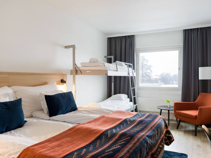 A hotel room with a bunk bed, a single bed, a small table with a lamp, a chair, and a window with curtains.