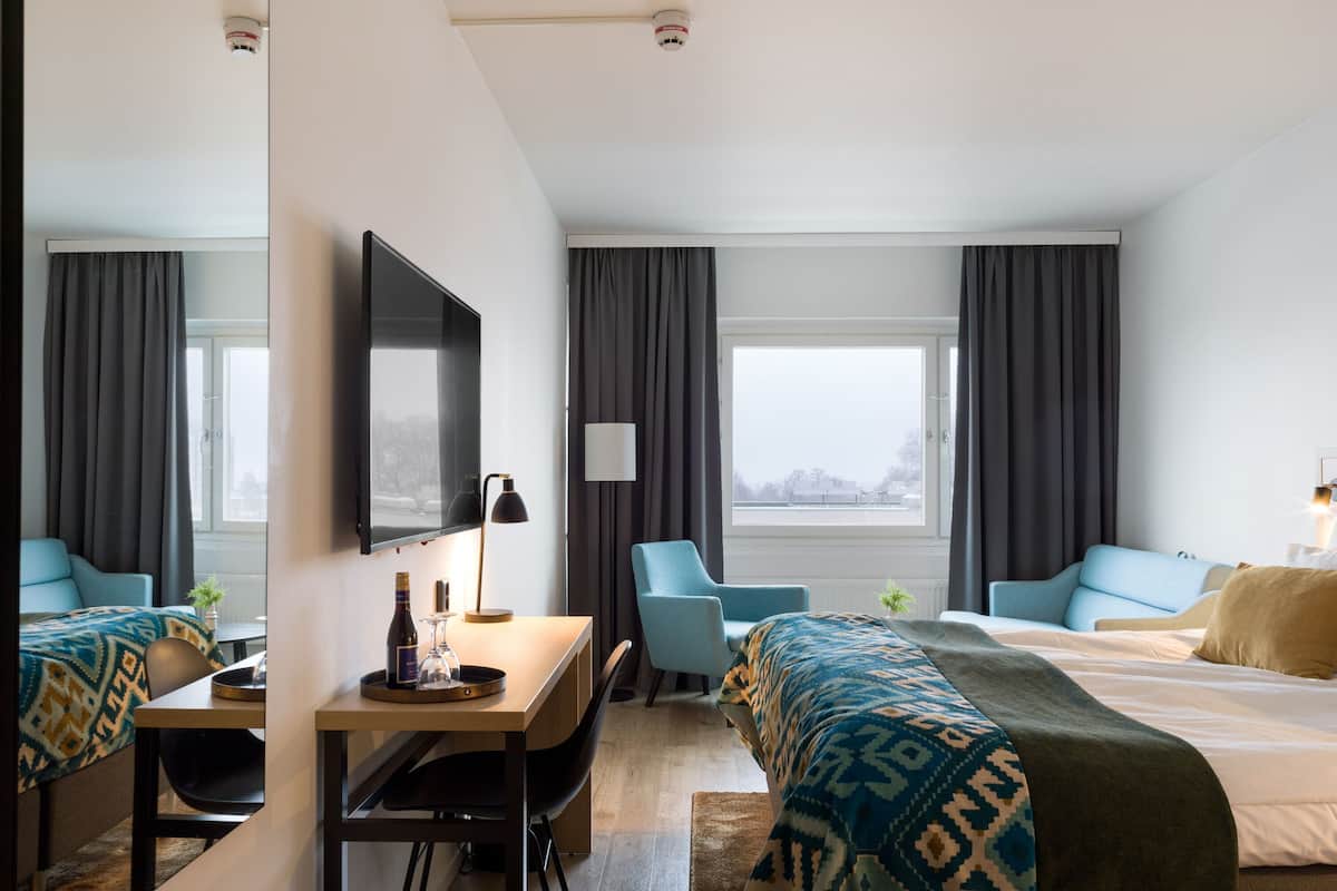 Hotellihuone, jossa on sänky, työpöytä tuolilla, televisio ja ikkuna, jossa on verhot.