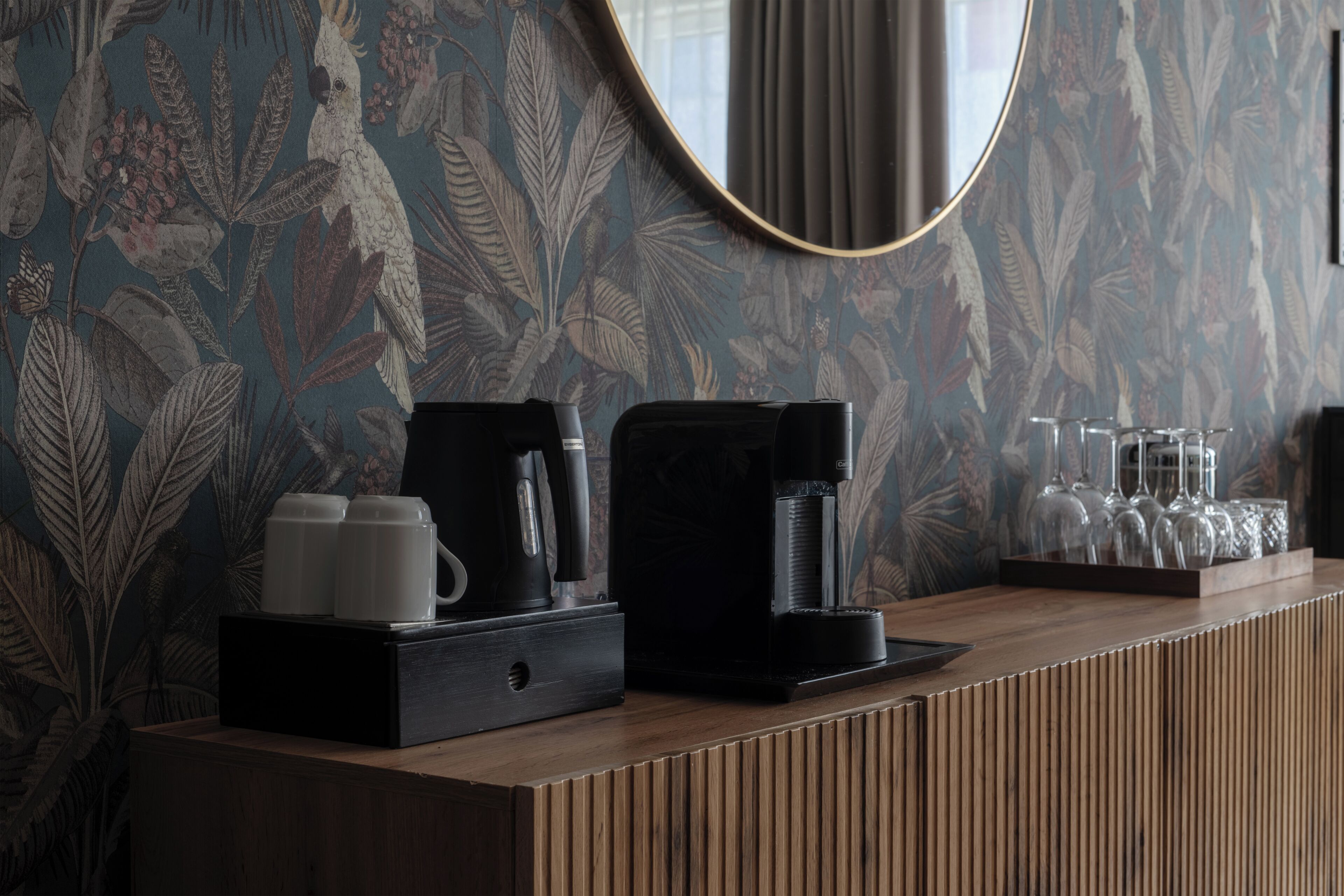 Un mostrador de recepción con una cafetera, tazas y vasos, frente a un papel tapiz floral y un espejo redondo.