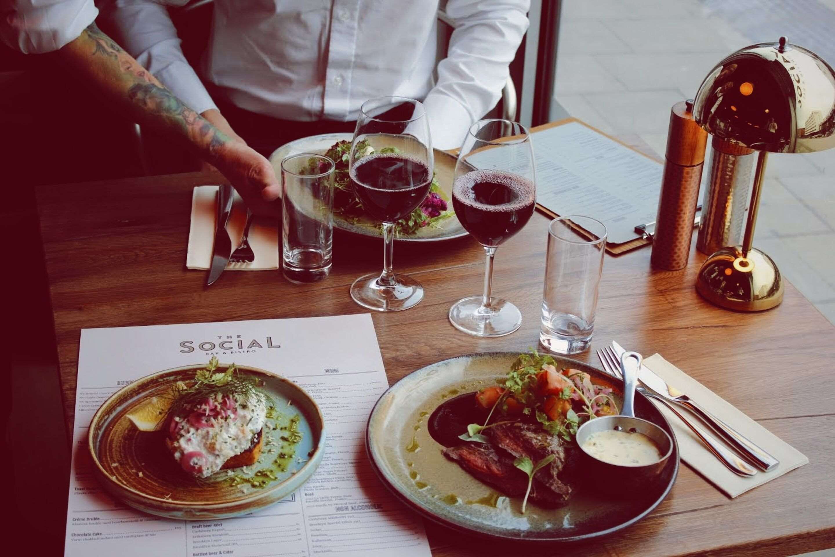 Una mesa de comedor con un menú, dos platos de comida, copas de vino y cubiertos.