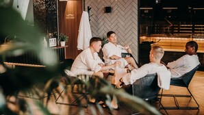 A group of people in white robes seated on lounge chairs in a modern, well-lit indoor setting with a herringbone-patterned wall and a wooden door labeled "Gentlemen."