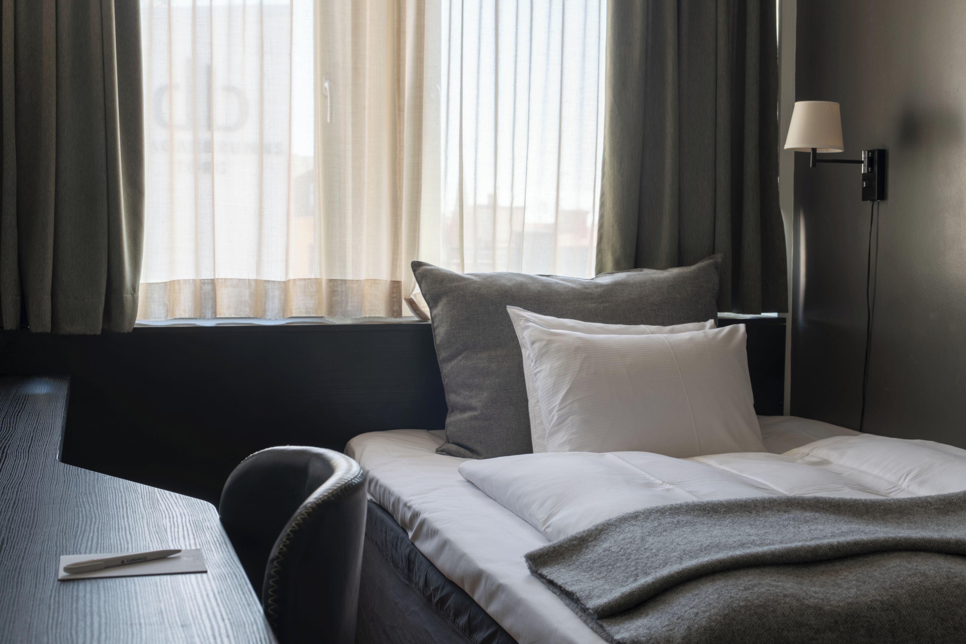 A hotel room with a bed, a desk, a lamp, and a window with curtains.