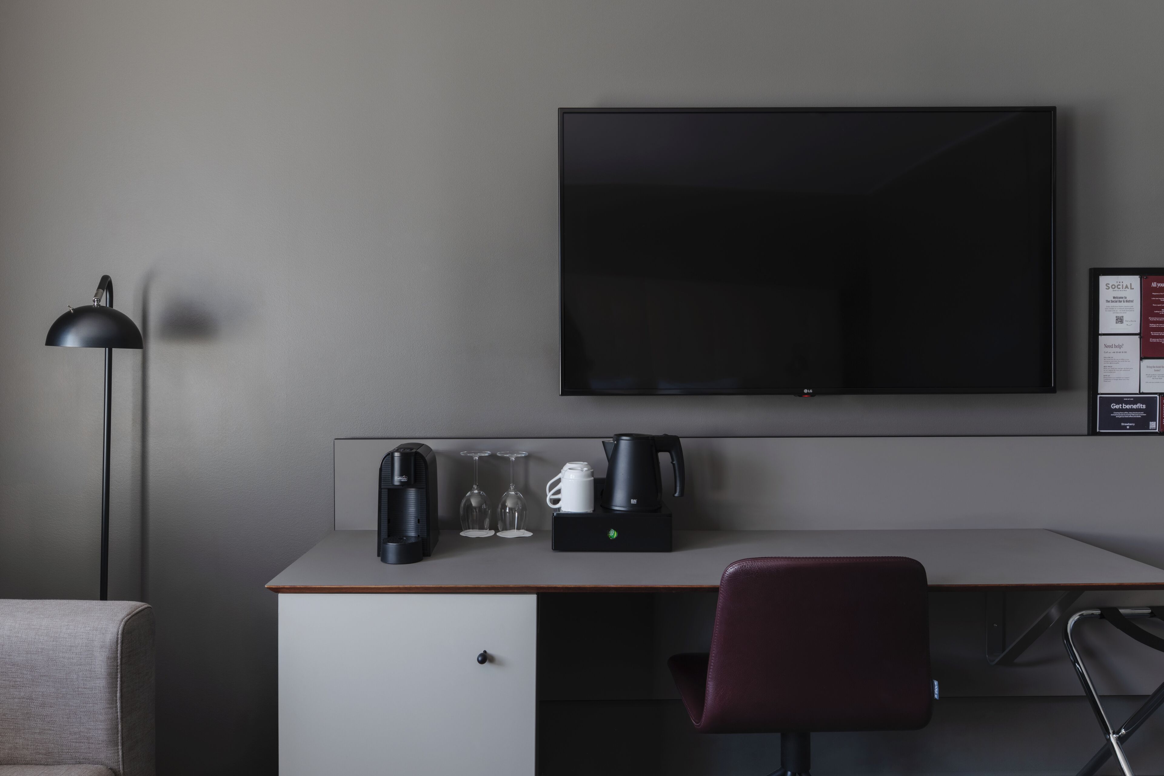 A hotel room with a desk, a television, a coffee maker, a cup, and a chair.