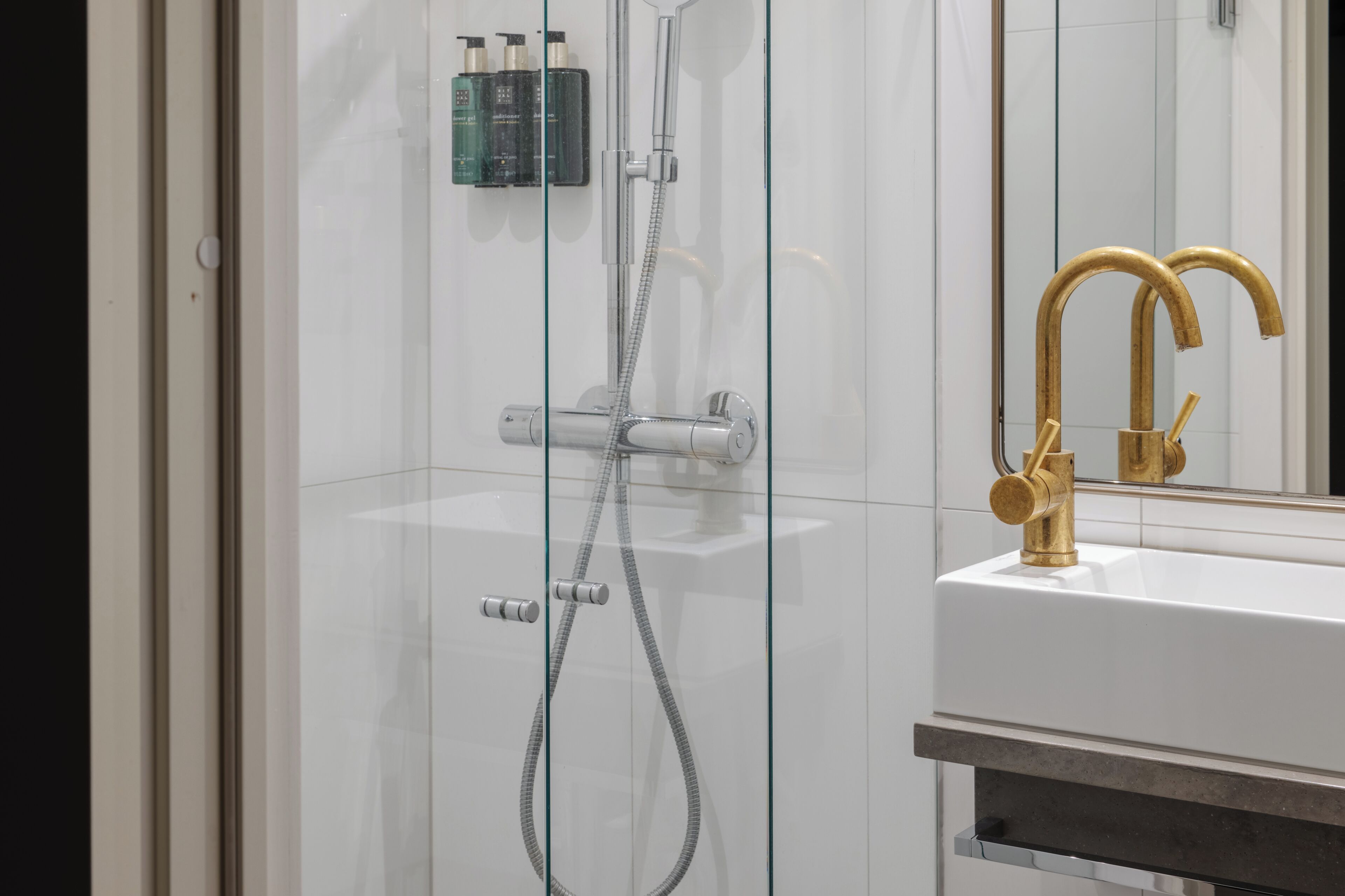 A modern bathroom with a glass-enclosed shower, a white sink with gold fixtures, and a mirror reflecting the space.