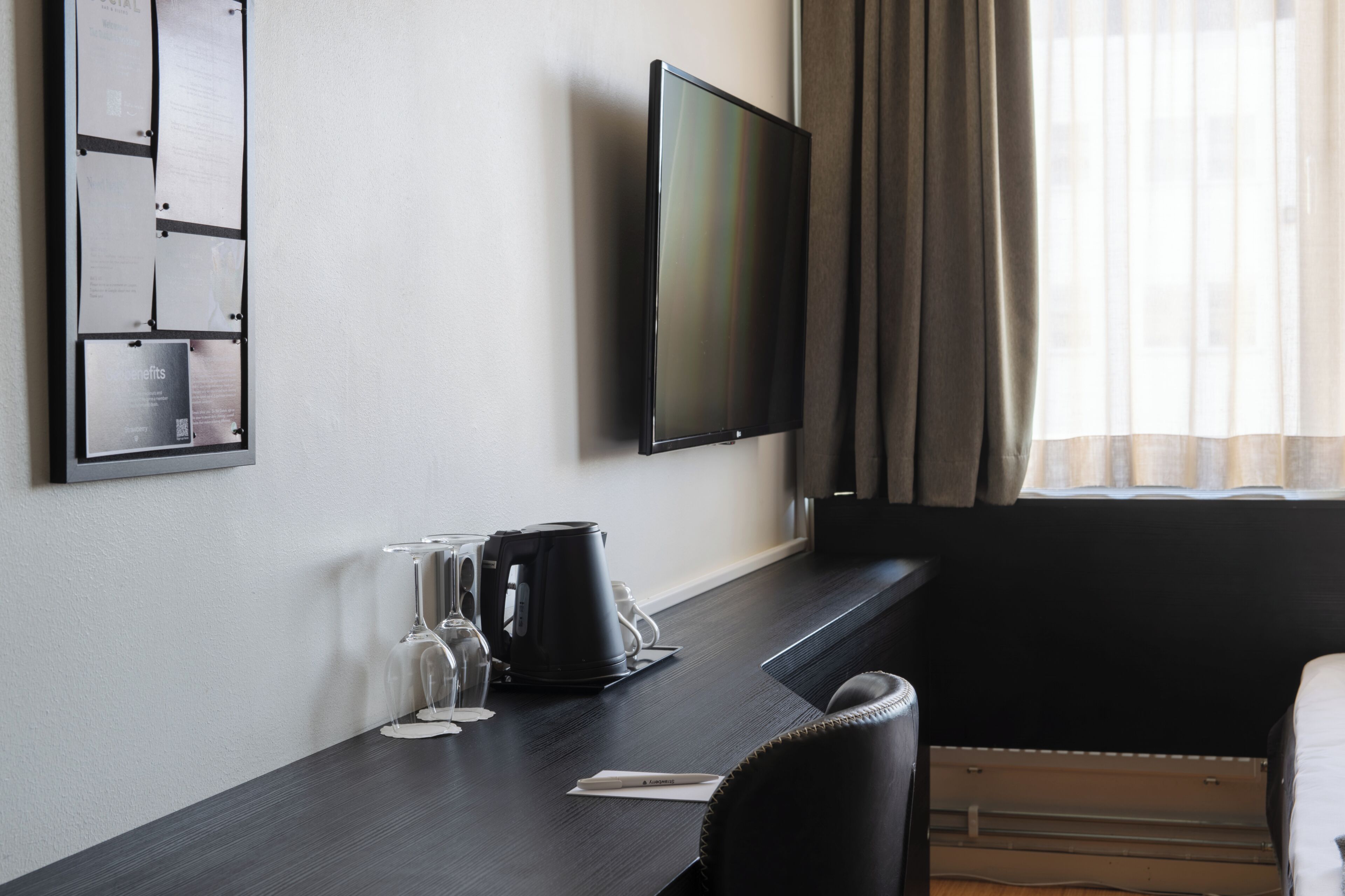 A hotel room with a dark wooden desk, a flat-screen TV, a coffee maker, wine glasses, and a comfortable chair.