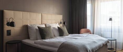A modern hotel room with a large bed, two bedside tables, a chair, and a window with curtains.