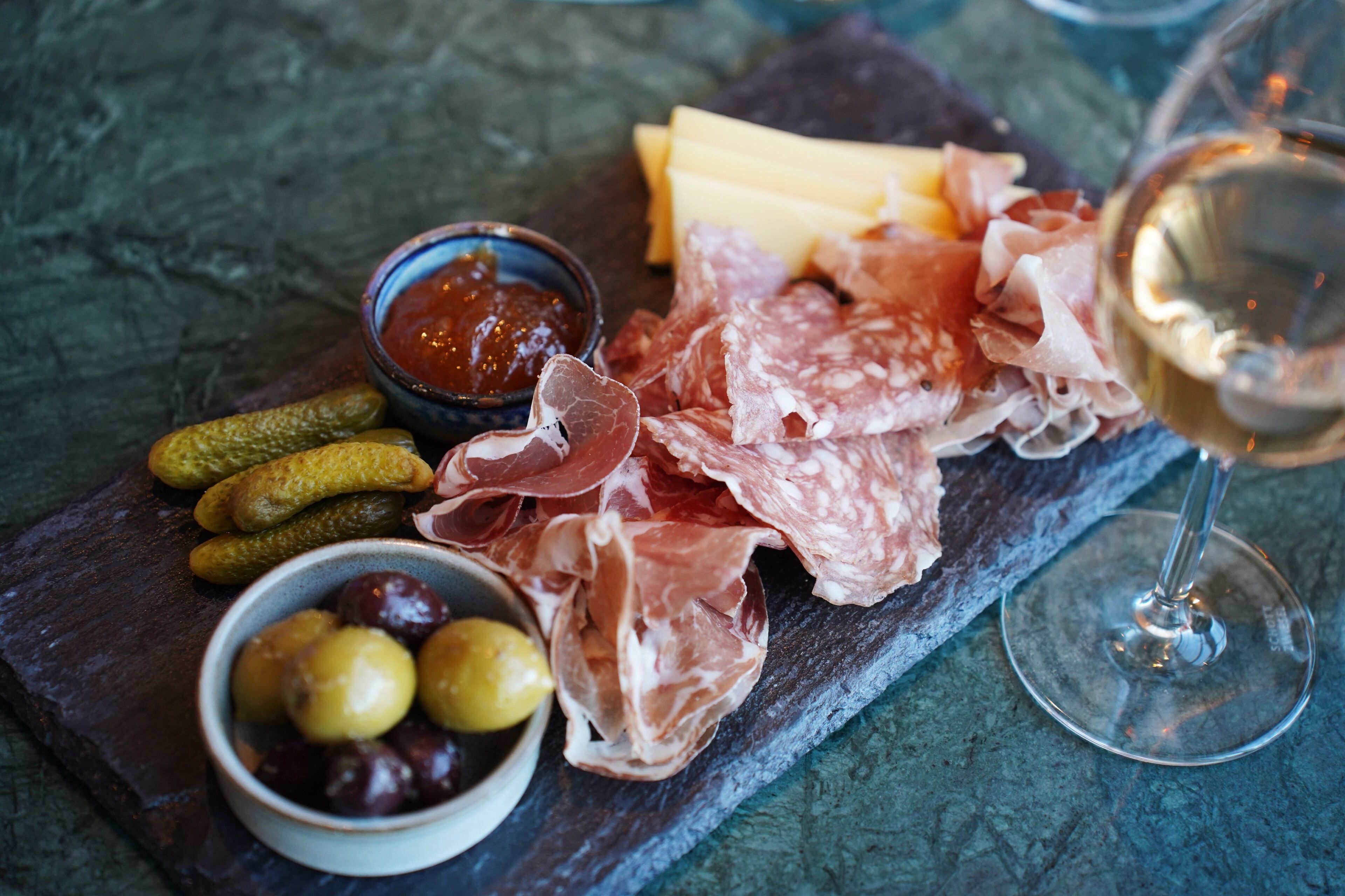 Una tabla de embutidos con diversos tipos de carne, quesos, aceitunas y pepinillos, acompañada de una copa de vino blanco.