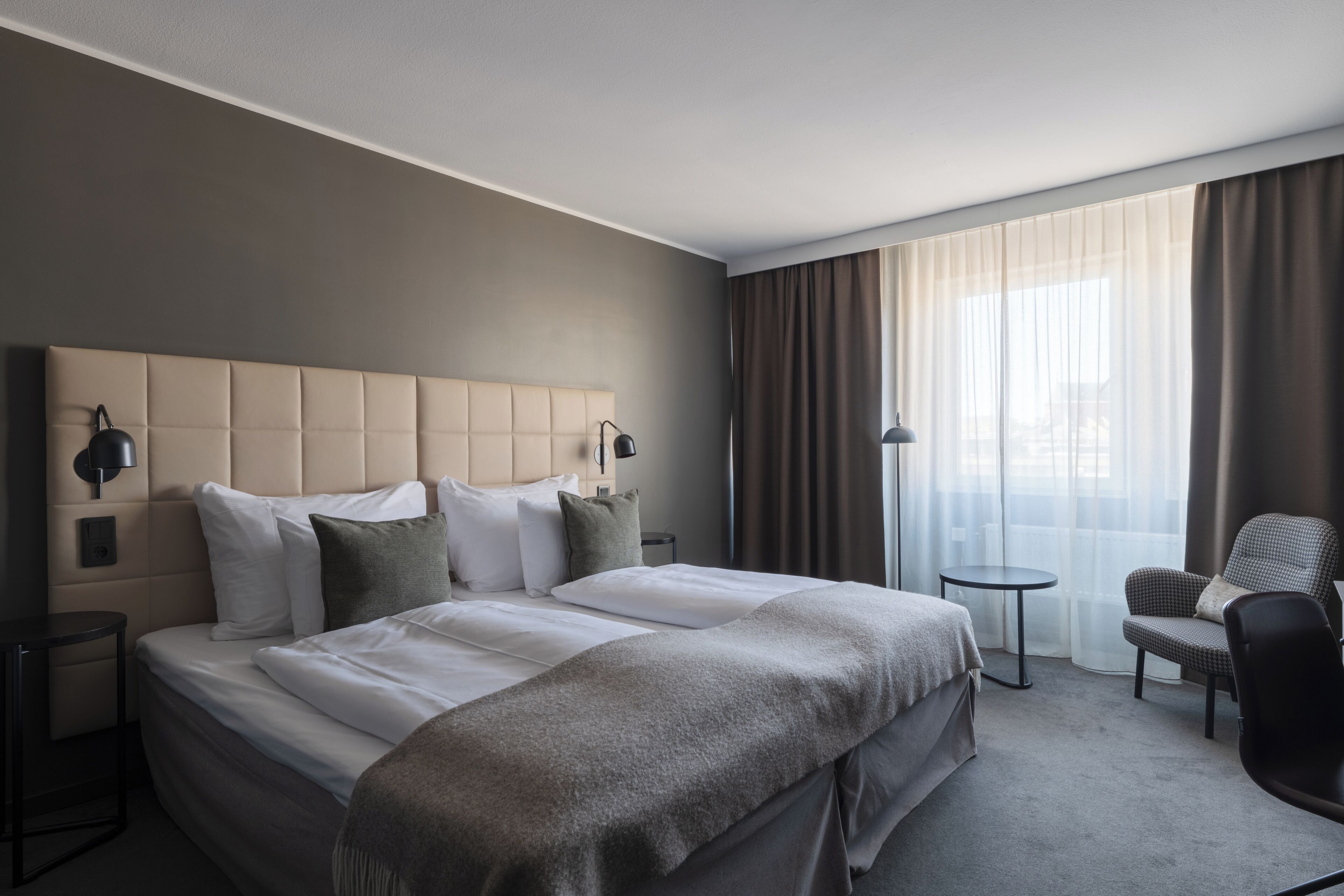 A hotel room with a large bed, a chair, a small table, and a window with curtains.