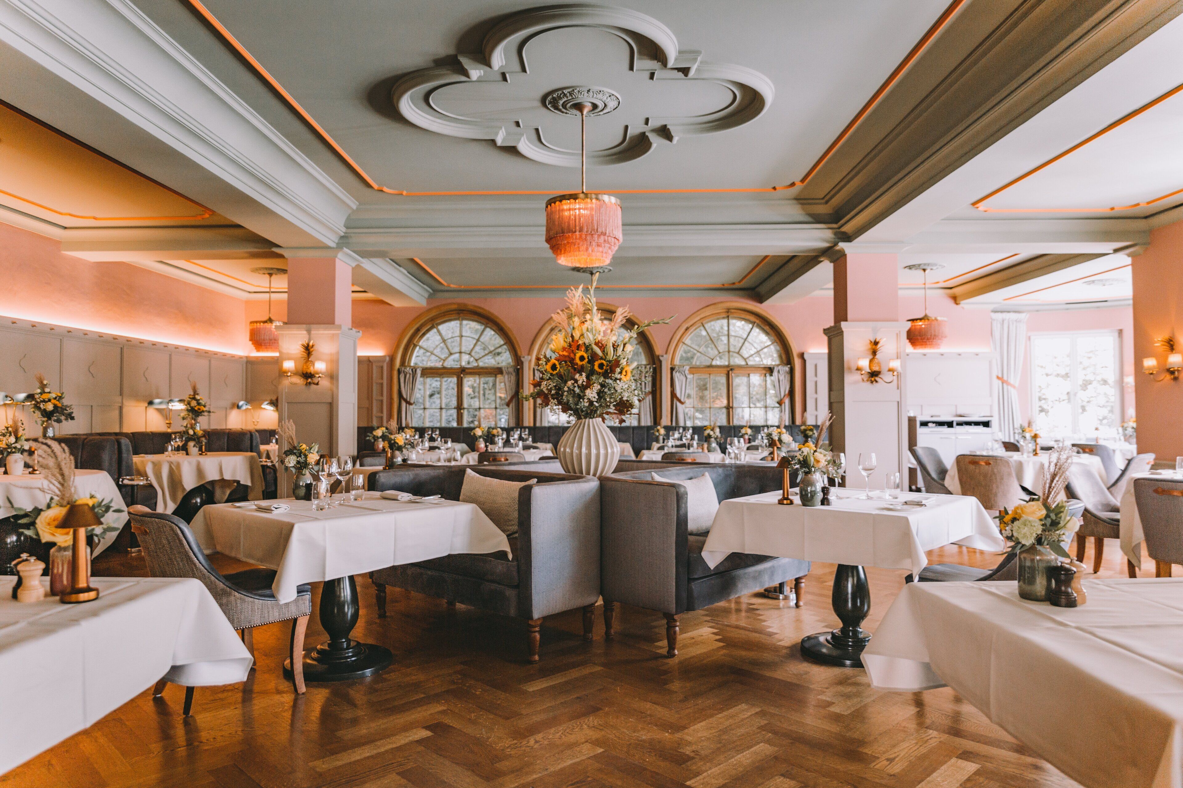 A restaurant with a classic interior, featuring wooden floors, white tablecloths, and a central chandelier.