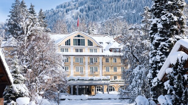Un hotel cubierto de nieve con un estilo arquitectónico clásico, que destaca por una entrada imponente y un paisaje de montañas al fondo.