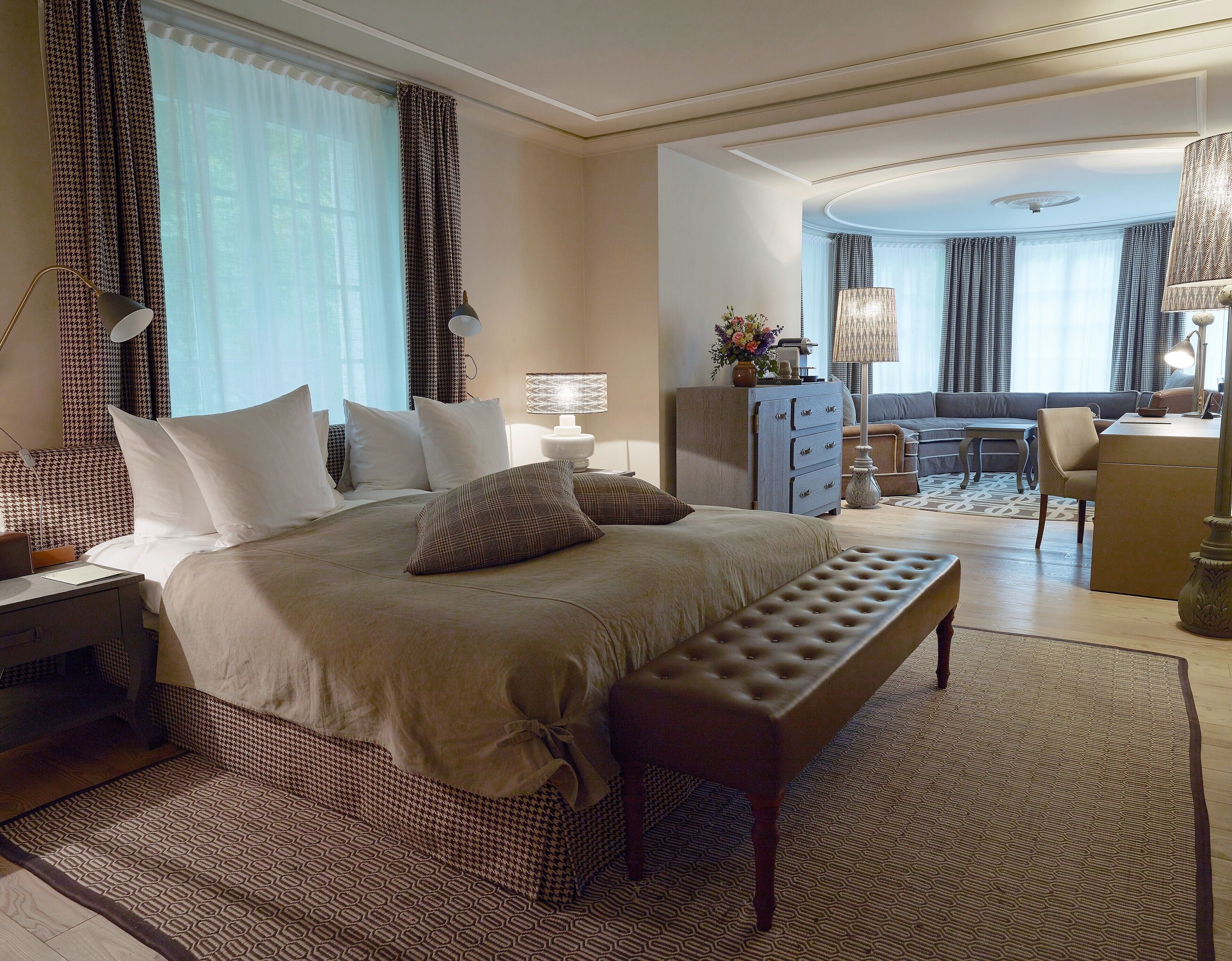 A hotel room with a large bed, a desk, a chair, a sofa, a dining table, and a chandelier.