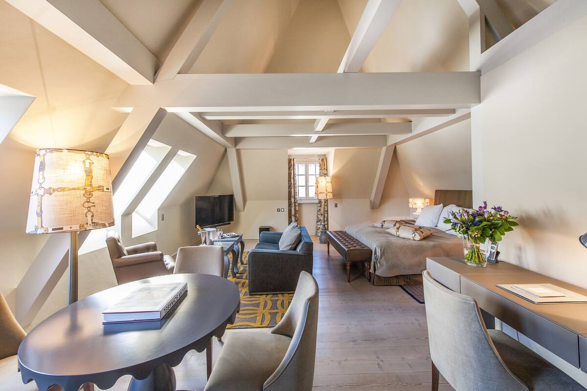 Un dormitorio tipo loft moderno con techo inclinado, una cama grande, una zona de estar con sofá y sillones, una mesa de comedor y un televisor.