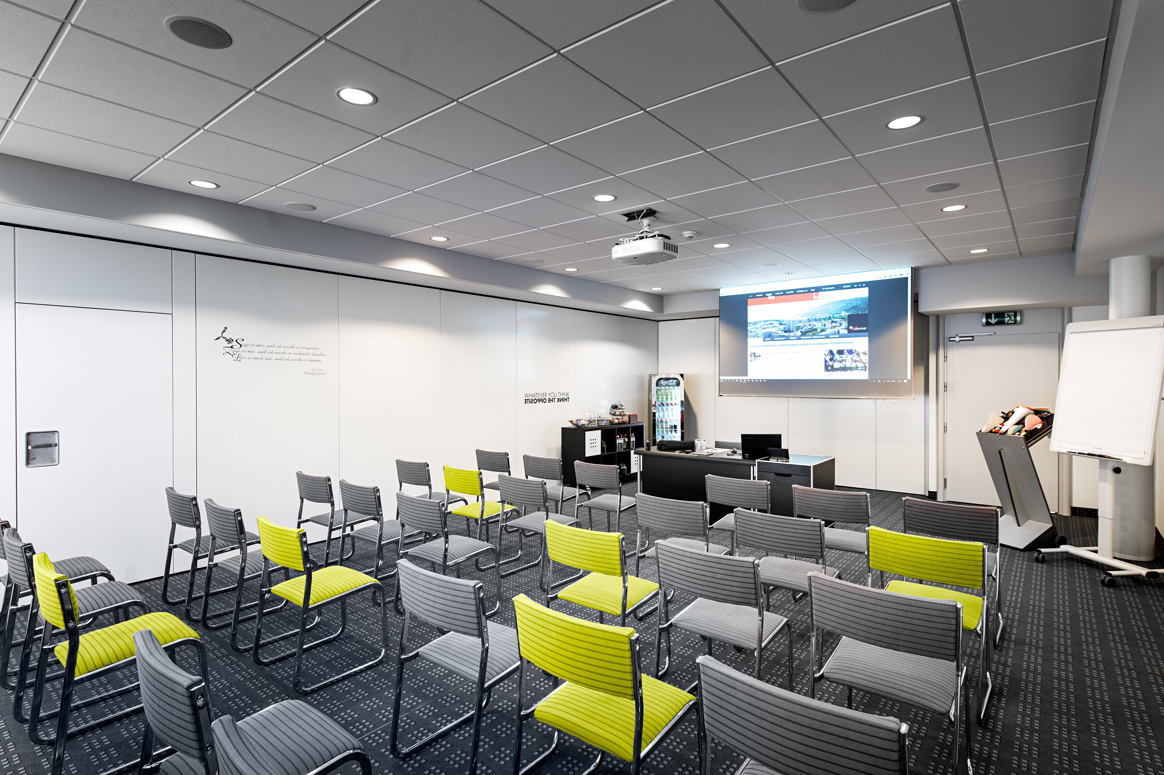Ein Konferenzraum mit Reihen von Stühlen, einer Projektionsleinwand, einer Whiteboard und einem Empfangstresen.