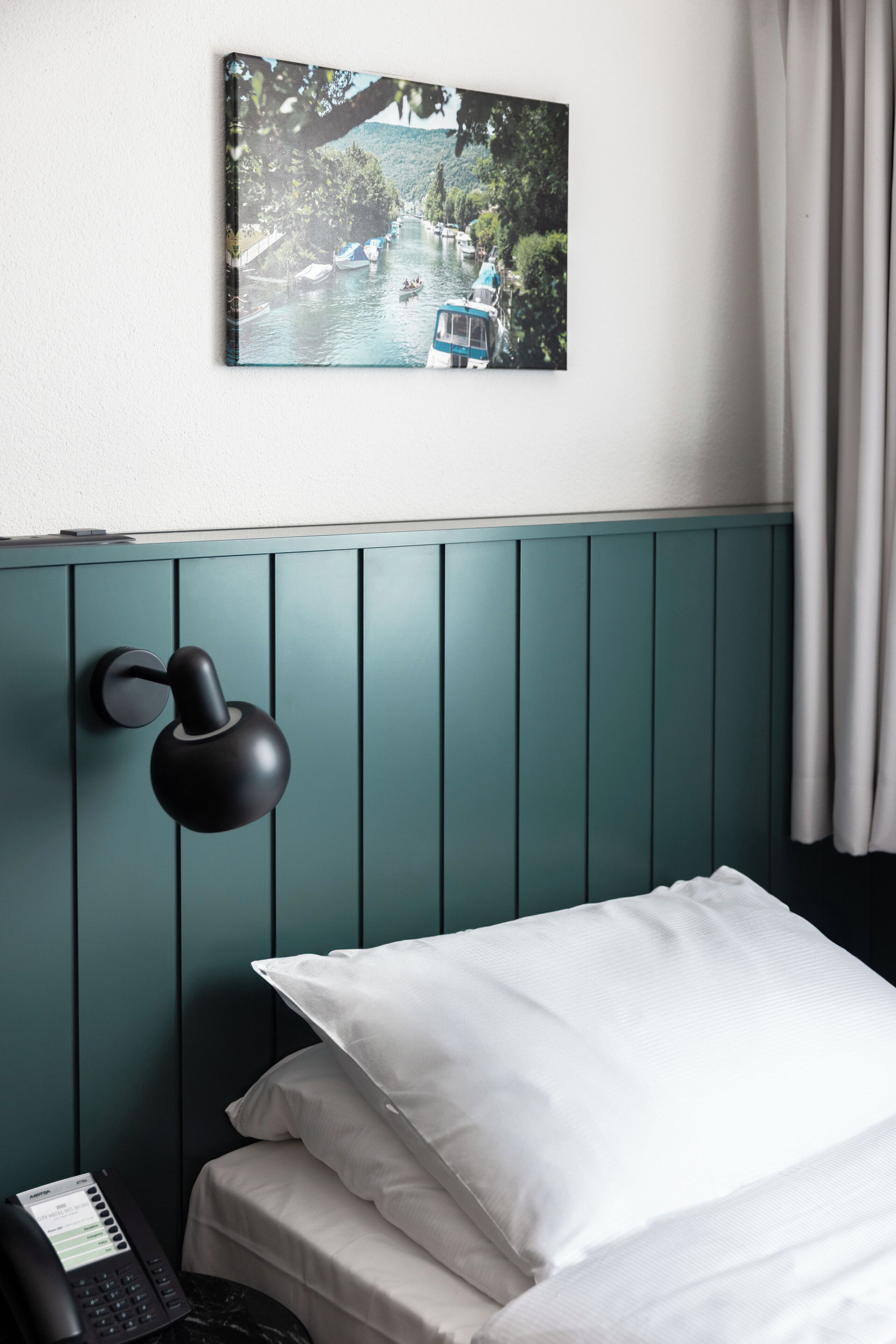 A hotel room with a bed, a wall-mounted lamp, a telephone, and a framed picture of a river scene.