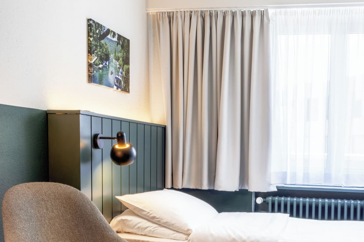 Ein modernes Hotelzimmer mit Holzboden, einem Bett mit weißen Leinen, einem grauen Sessel, einem kleinen Tisch mit Früchten, einer Wandlampe und einem Fenster mit Vorhängen.