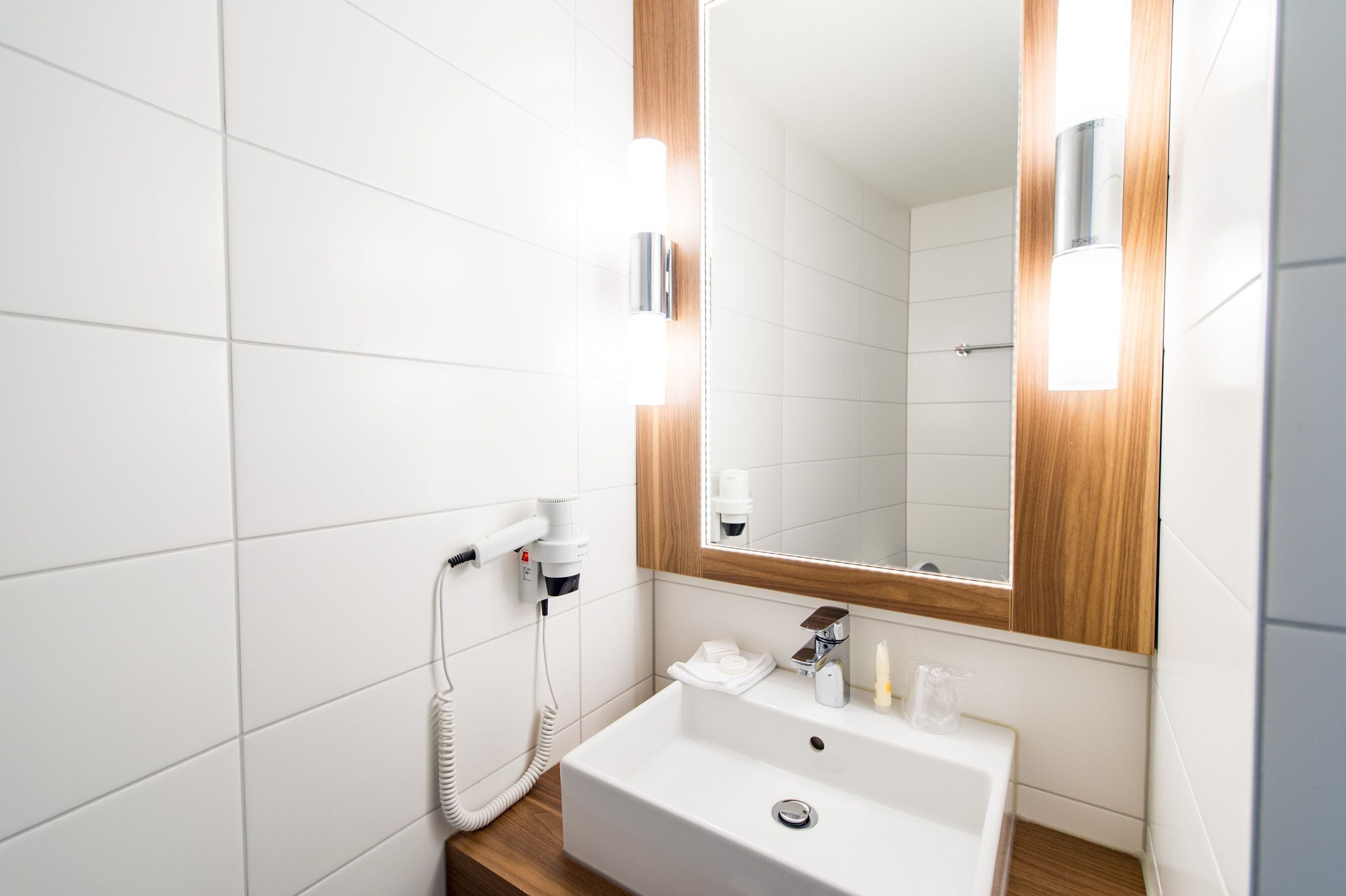 Une salle de bain moderne avec des carreaux blancs, un meuble-lavabo en bois, un grand miroir et un sèche-cheveux fixé au mur.