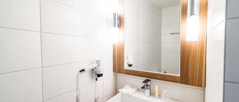 A modern bathroom with white tiles, a wooden vanity, a large mirror, and a hairdryer on the wall.
