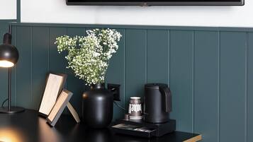 A modern room with a flat-screen TV mounted on the wall, a wooden desk with a lamp, a vase with flowers, a book, and a coffee machine.
