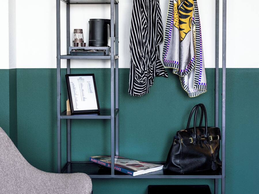 Una habitación con un sillón gris, un bolso de cuero negro, un perchero con varias prendas, una estantería con libros y un certificado enmarcado, y una pequeña caja fuerte.
