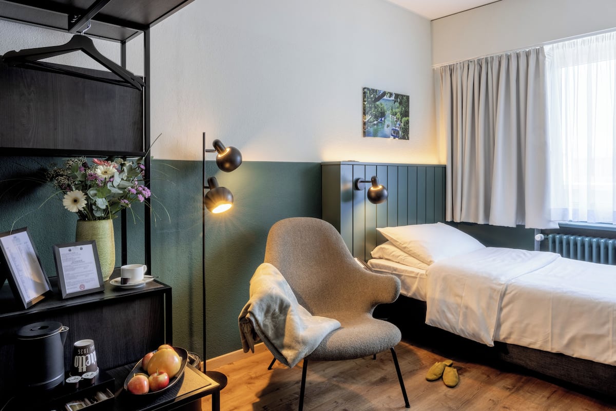 Ein Hotelzimmer mit Bett, Sessel, Schreibtisch mit Lampe, Bilderrahmen, Blumen in einer Vase und Tablett mit Äpfeln und Kaffeemaschine.