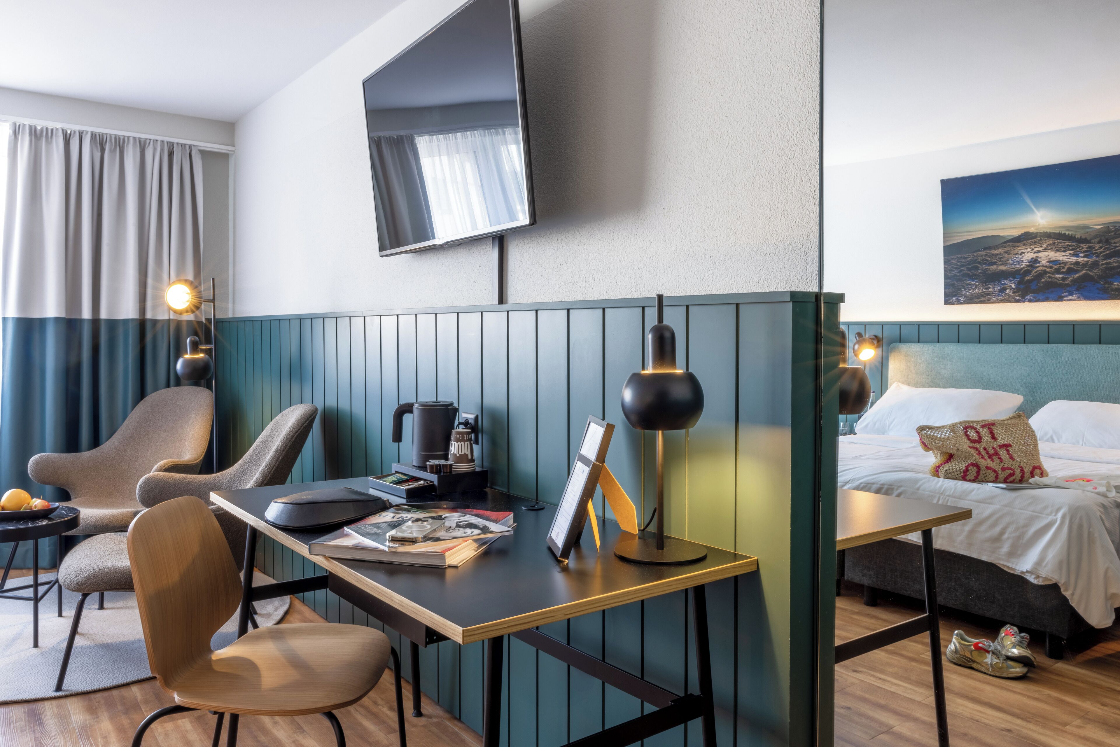 Ein Hotelzimmer mit Schreibtisch, Stühlen, einem Bett, einem Fernseher und einem Bild an der Wand.