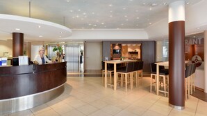 Un moderno vestíbulo de hotel con mostrador de recepción, zona de comedor con mesas y sillas de madera, y cocina a la vista al fondo.