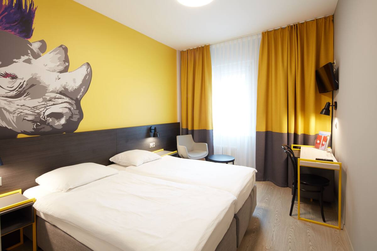 A hotel room with two beds, a desk, a chair, a TV, and a large wall mural.