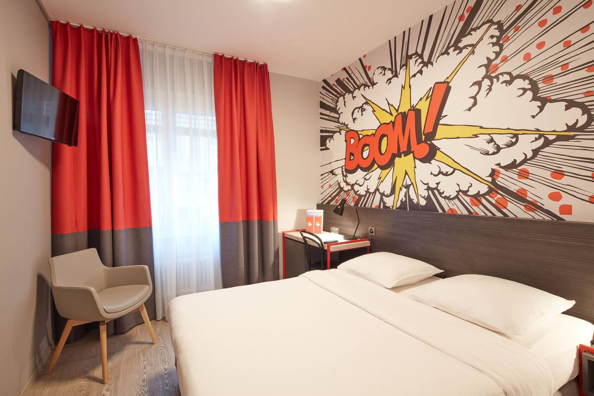 A hotel room with a bed, a desk, a chair, a TV, and a wall with a comic book style explosion design.