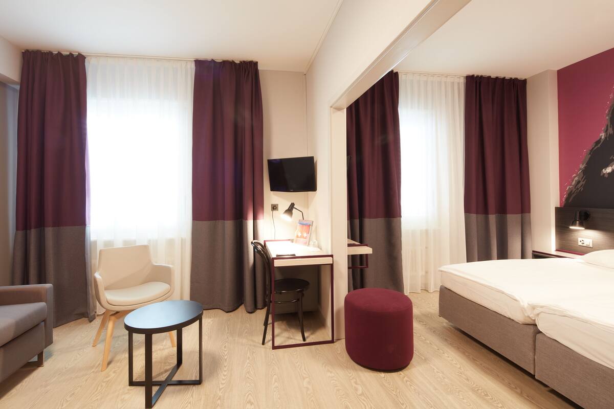 A hotel room with a bed, a desk with a chair, a small table, and a TV mounted on the wall.