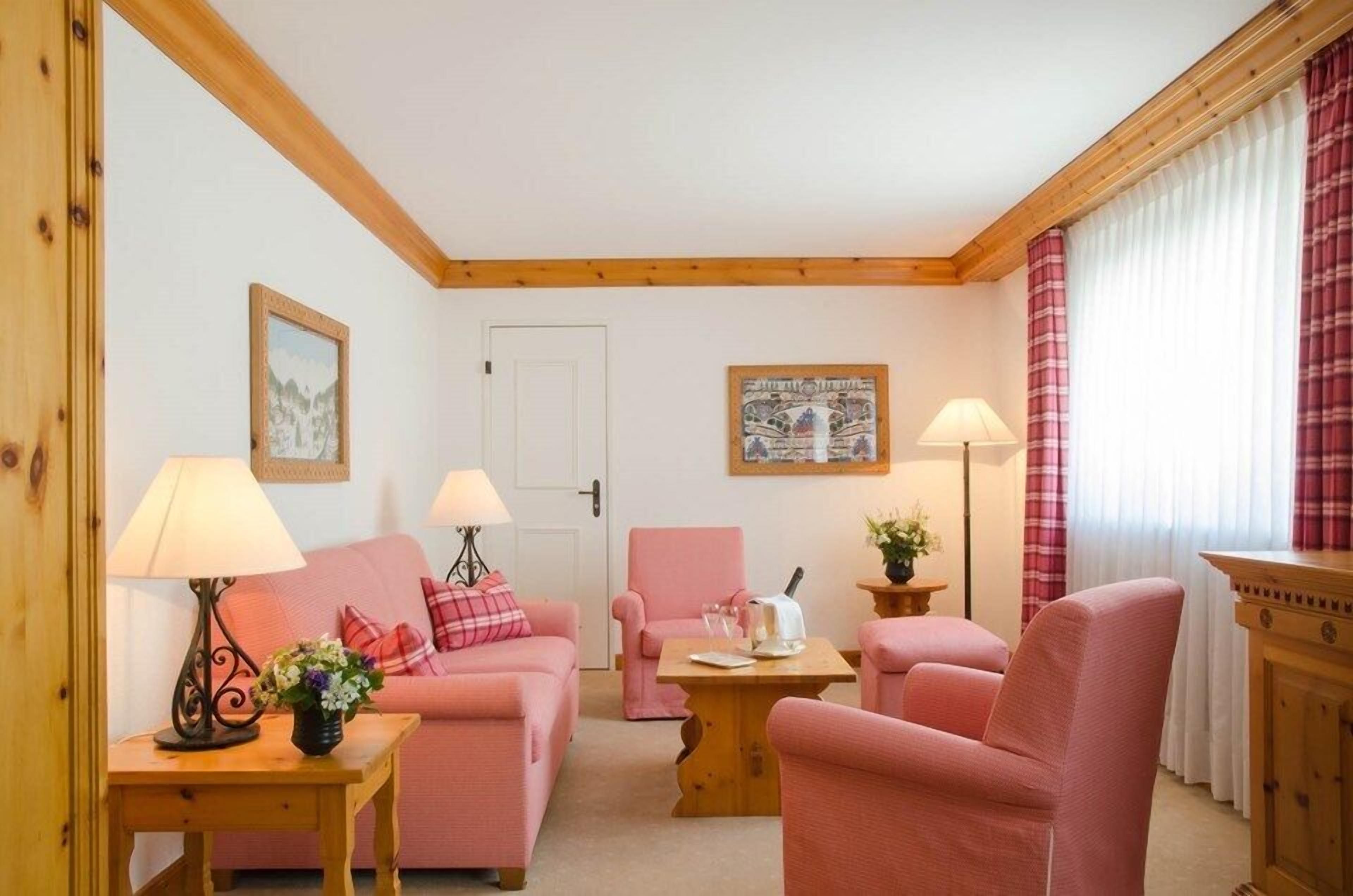 Ett mysigt vardagsrum med en rosa soffa, tillhörande fåtöljer och ett soffbord i trä. Det står två lampor på sidoborden, en inramad tavla hänger på väggen och en dörr syns i bakgrunden.