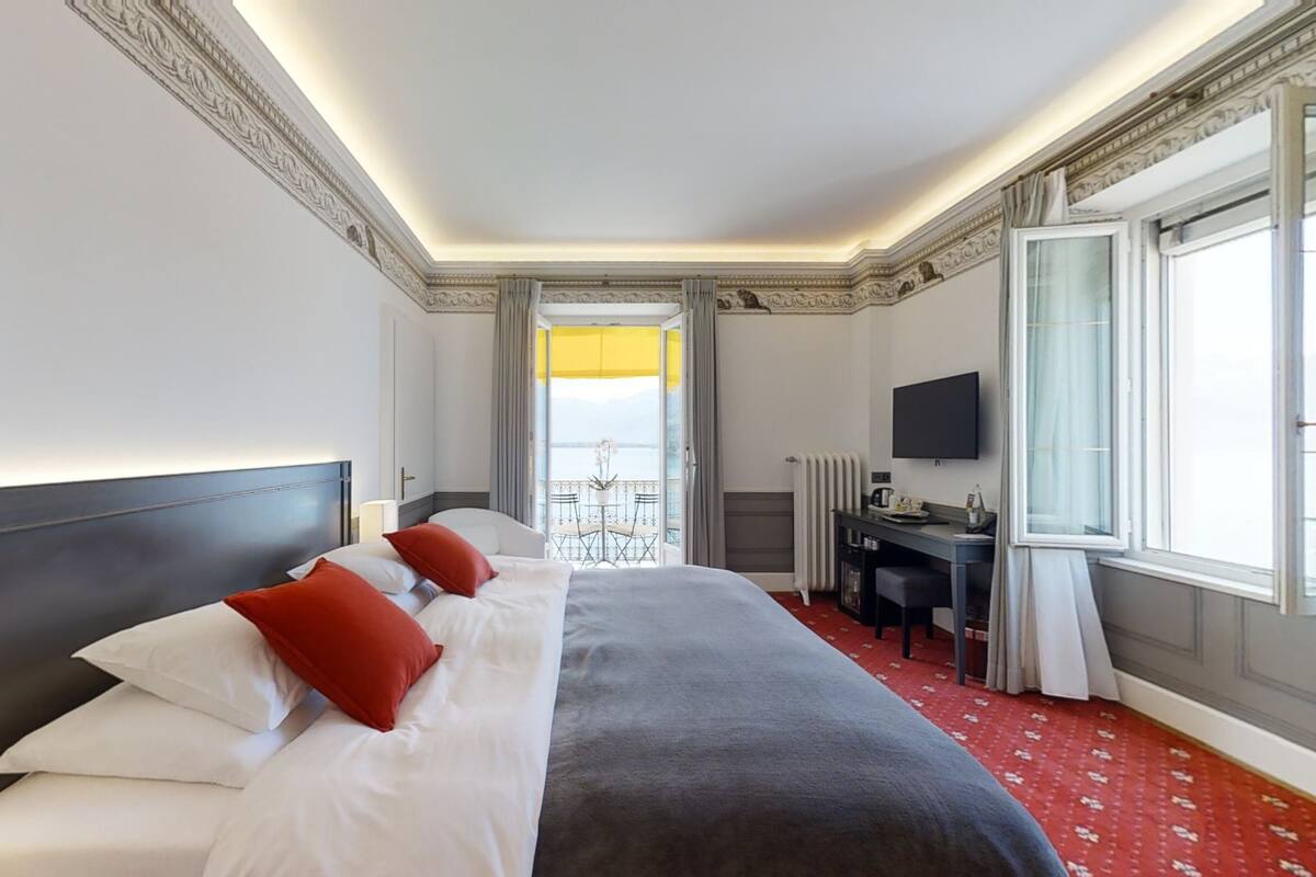 A hotel room with a large bed, a desk, a chair, a TV, and a balcony with a view.