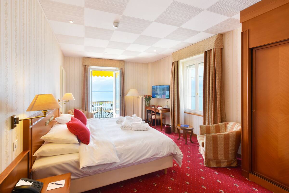 A hotel room with a large bed, a desk, a chair, a TV, and a balcony with a view.
