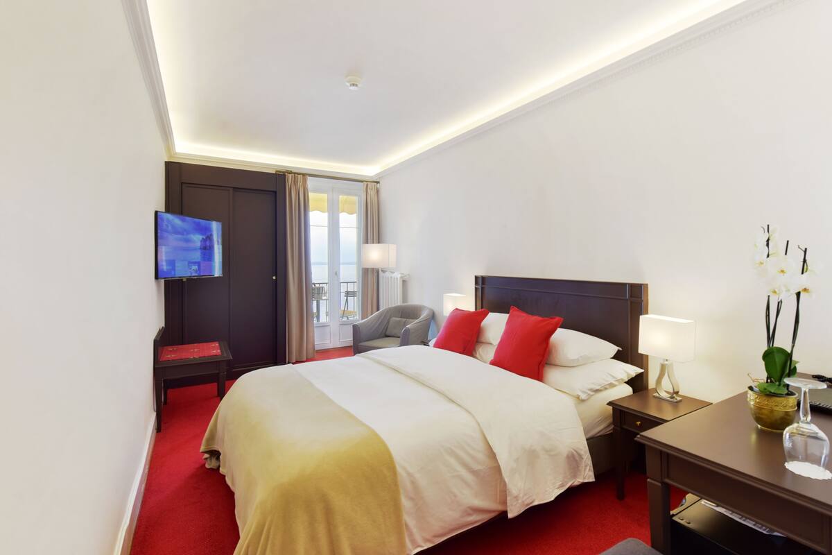 A hotel room with a large bed, a TV, a desk with a lamp, and a view of the outdoors.