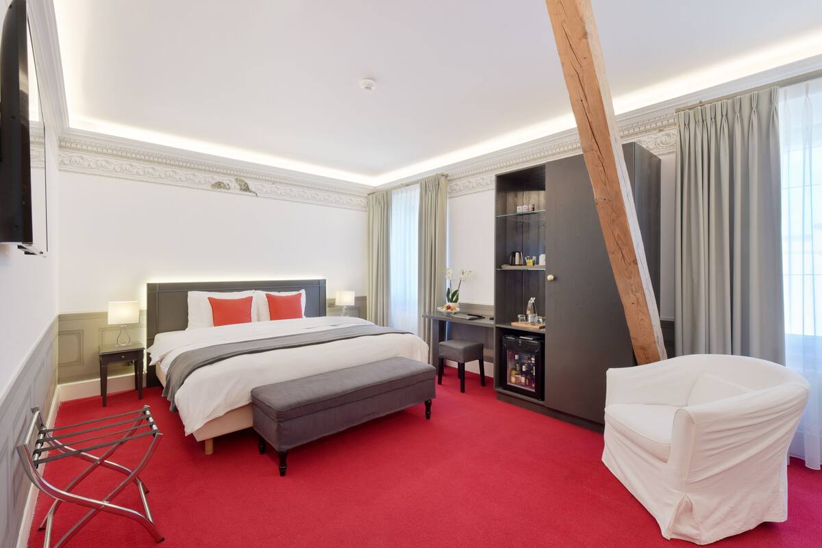 A hotel room with a bed, a desk, a chair, a wardrobe, and a red carpet.
