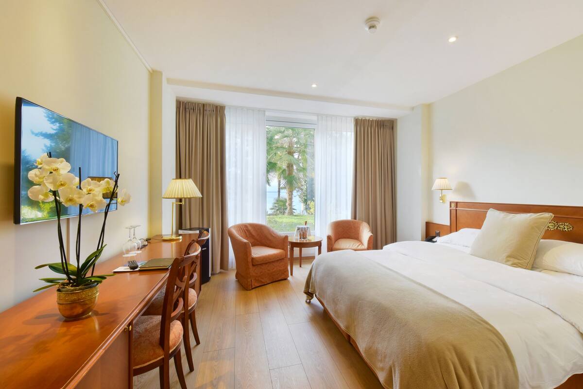 A hotel room with a bed, two chairs, a desk with a laptop, a vase with flowers, and a window with curtains.