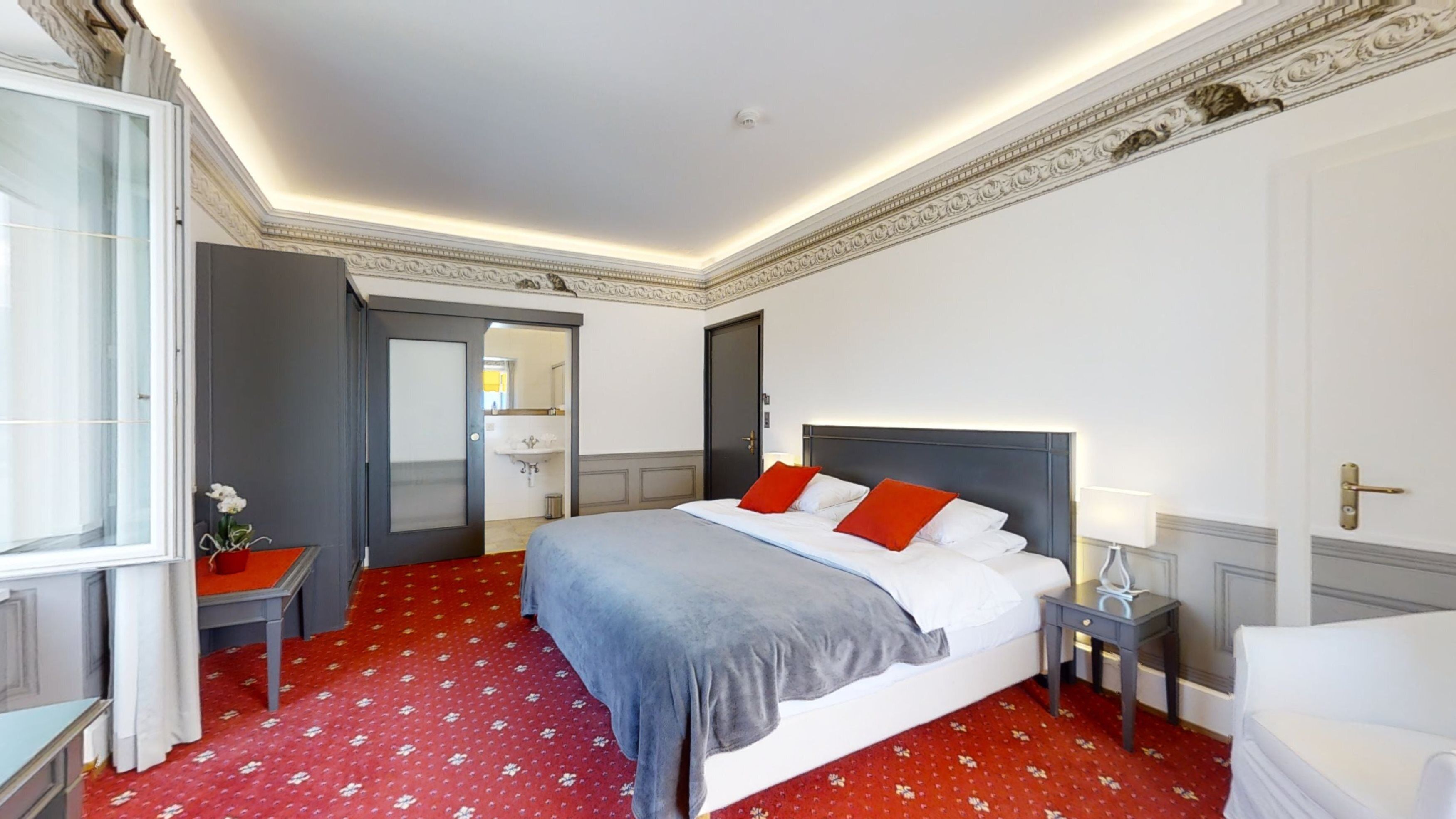 Une chambre d’hôtel avec un grand lit, une table de chevet, une commode, une chaise et une fenêtre avec des rideaux.