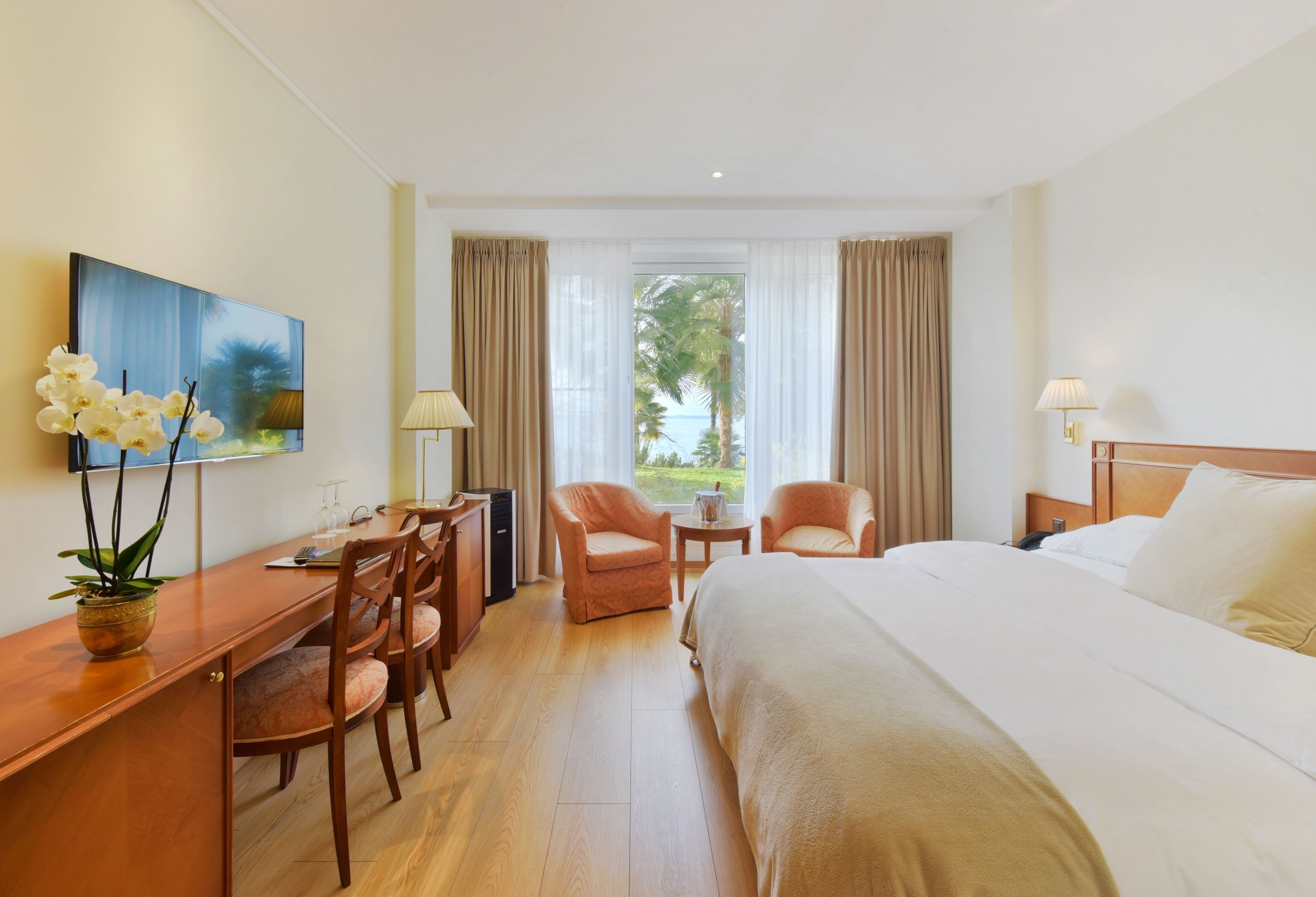 A hotel room with a bed, two armchairs, a desk, and a view of palm trees through the window.