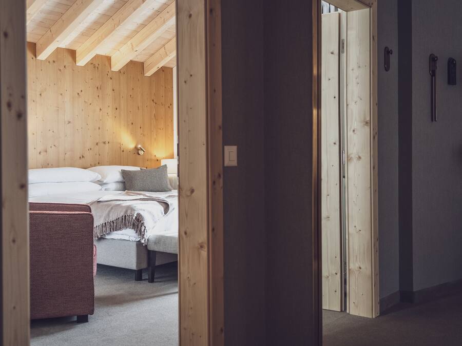 Una camera da letto con soffitto in legno, un letto con lenzuola bianche, una panca intrecciata rossa e una porta con maniglia.