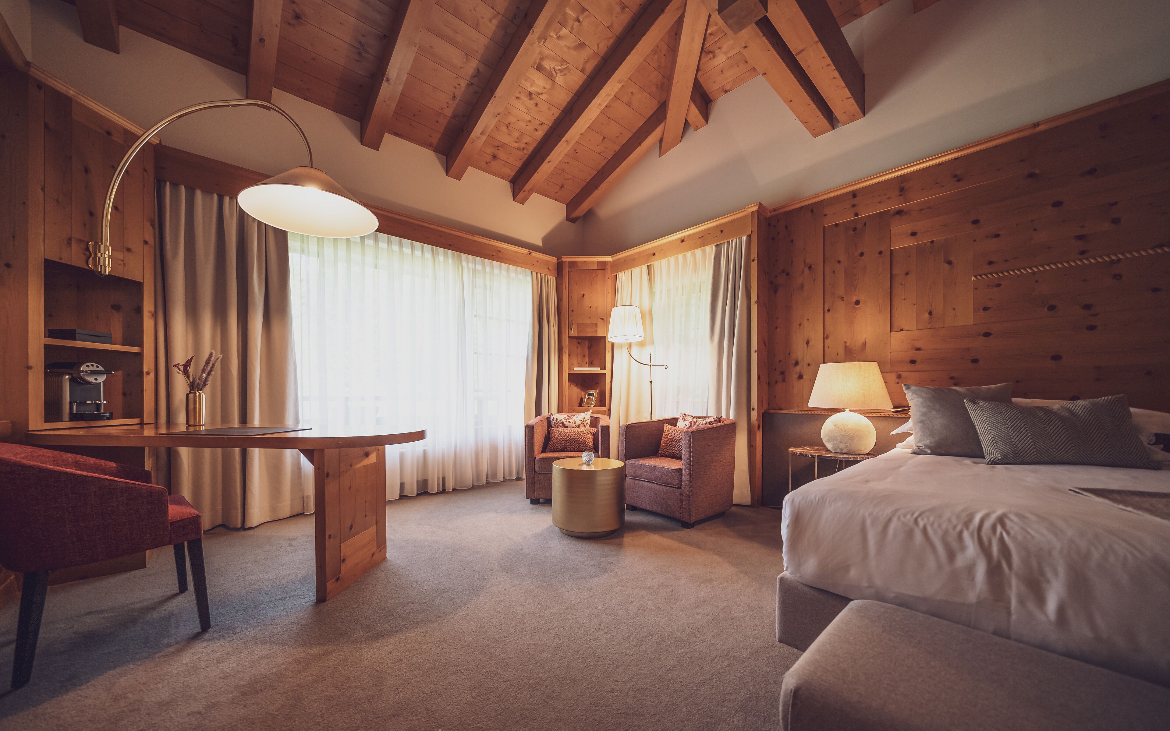 A cozy hotel room with a wooden ceiling, a bed, a desk, and two armchairs.
