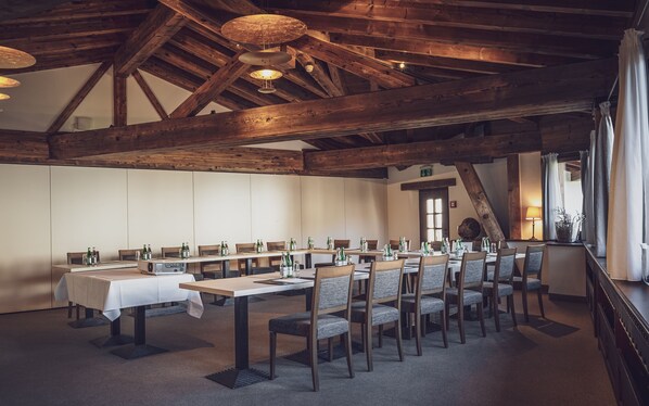 Una larga mesa de comedor con sillas, vigas de madera y lámparas colgantes en un interior rústico.