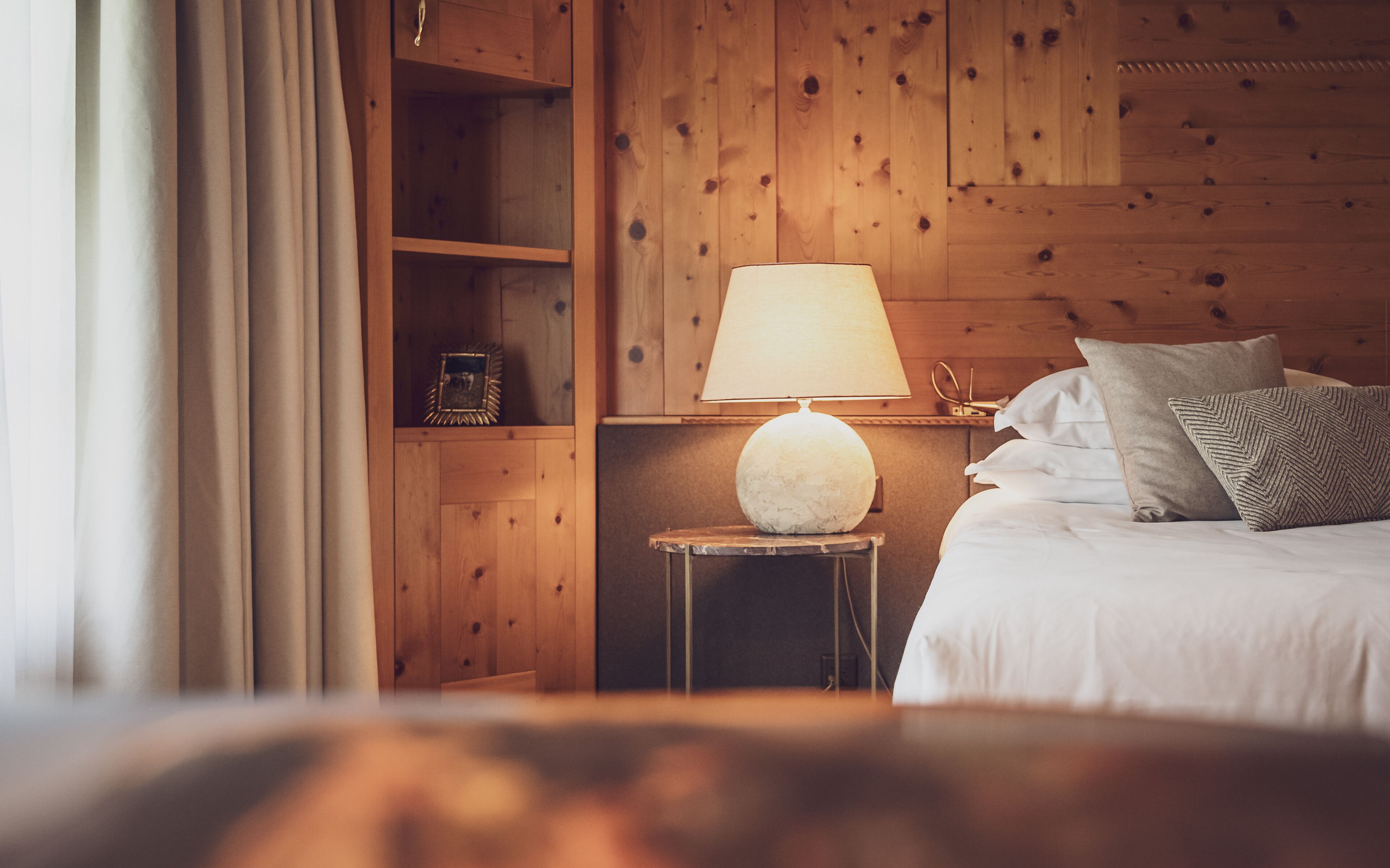 Ein Zimmer mit Holzboden, einem Bett, einem Nachttisch mit Lampe und einem Regal mit Bilderrahmen.