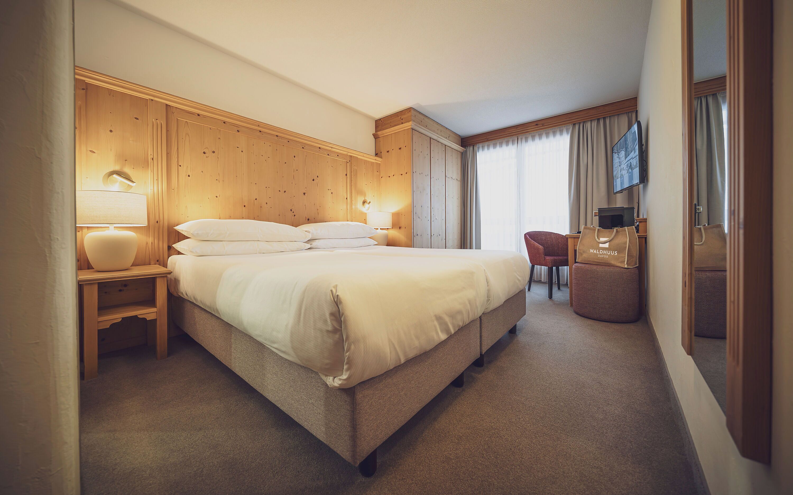 A hotel room with a bed, bedside lamp, wooden headboard, a desk with a chair, and a television.