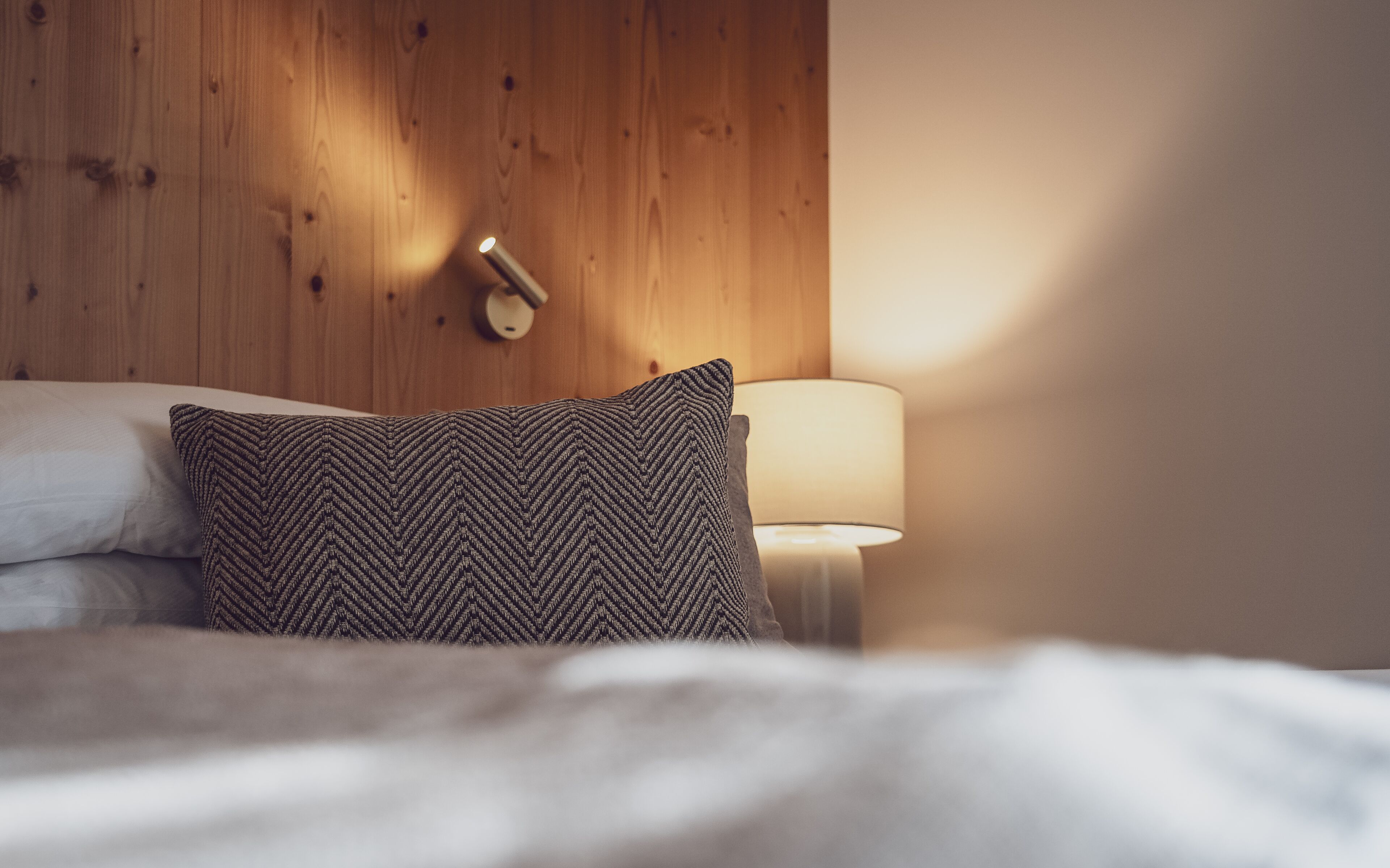 A neatly made bed with a patterned pillow and a bedside lamp against a wooden wall.