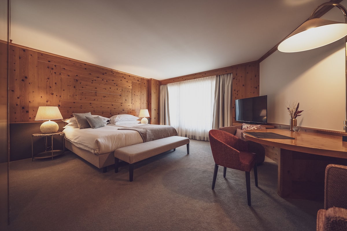 A hotel room with a wooden paneled wall, a bed with white linens, a desk with a chair, a television, and a large window with curtains.