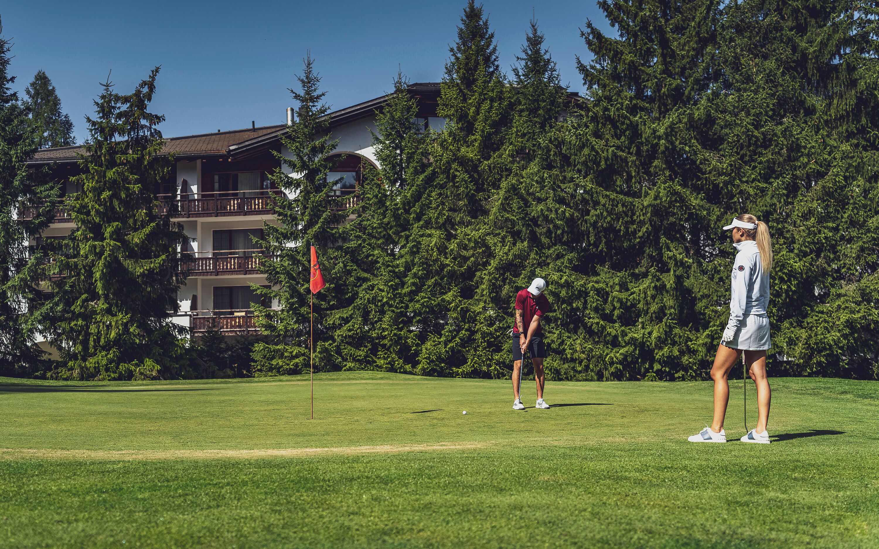 A golf course with a flag, trees, and a building with balconies in the background.