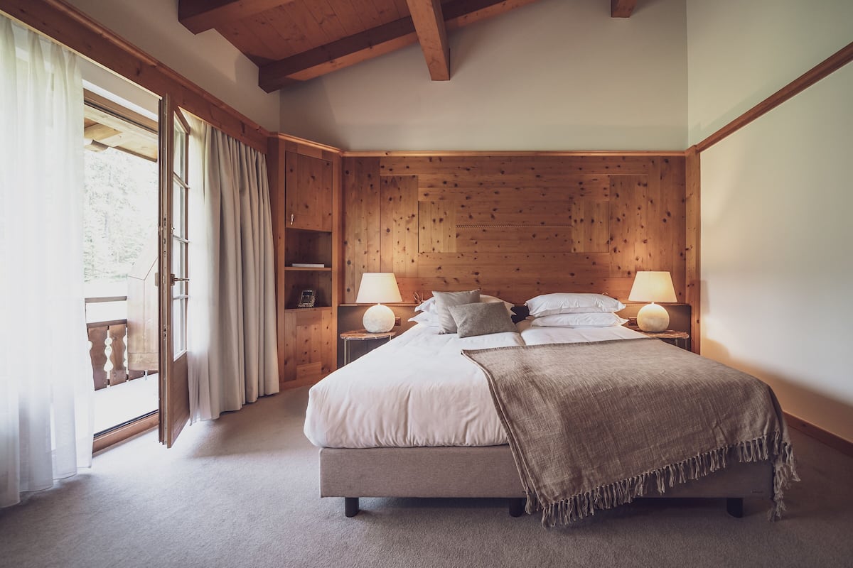 A bedroom with a large bed, wooden headboard, two bedside lamps, a nightstand, and a window with curtains.