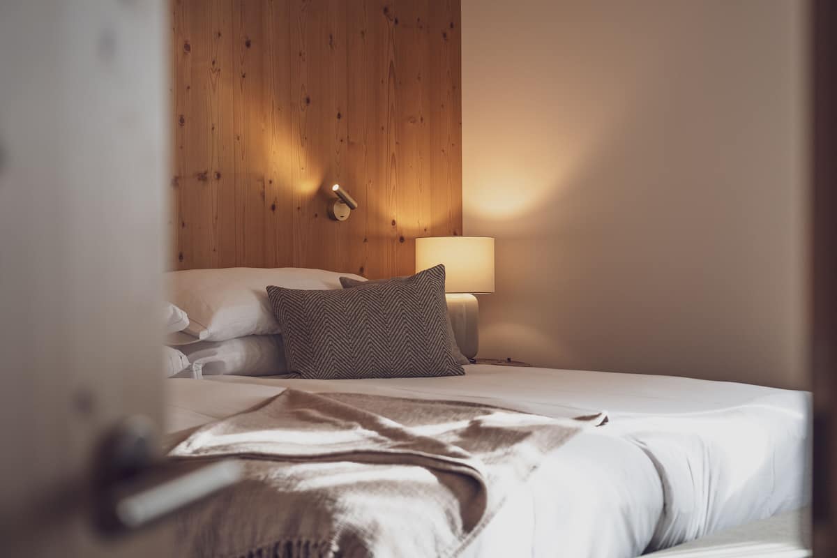 A neatly made bed with white linens and a brown throw blanket, a wooden headboard, a bedside lamp, and a wall-mounted light fixture.