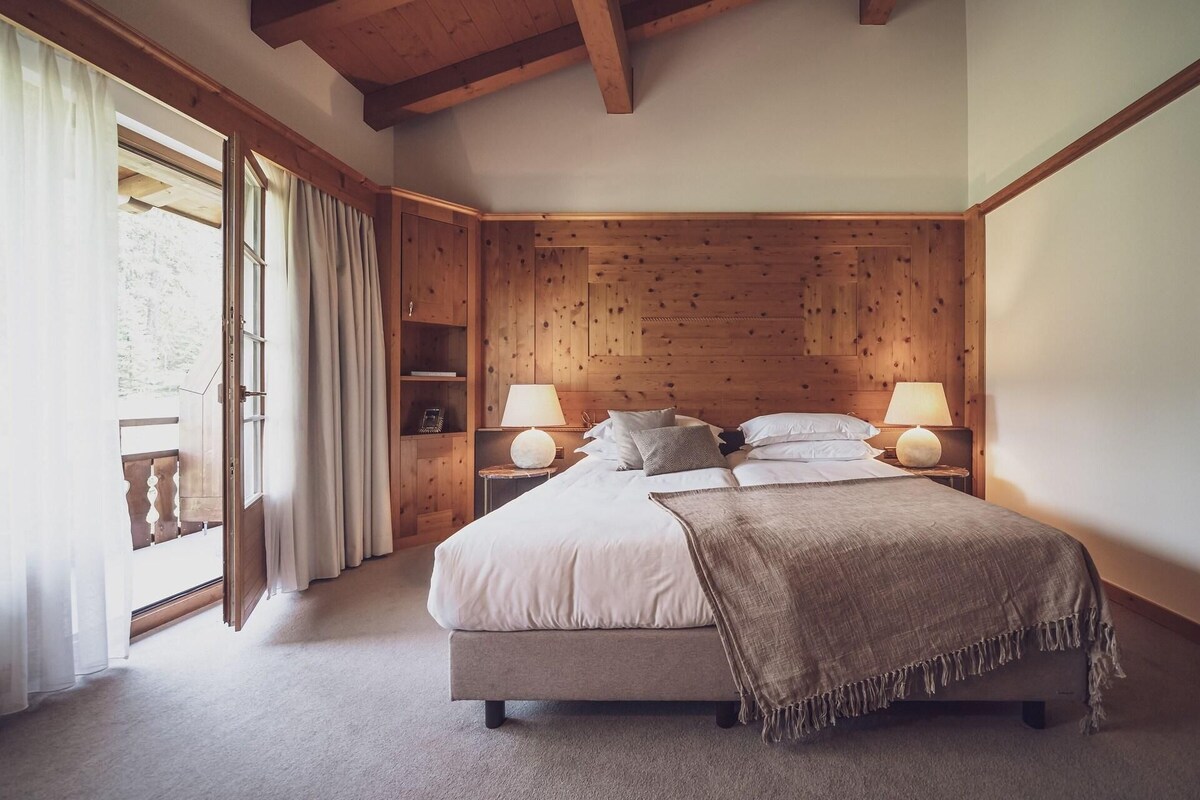 A bedroom with a large bed, wooden headboard, two bedside tables with lamps, a wooden wardrobe, and a window with curtains.