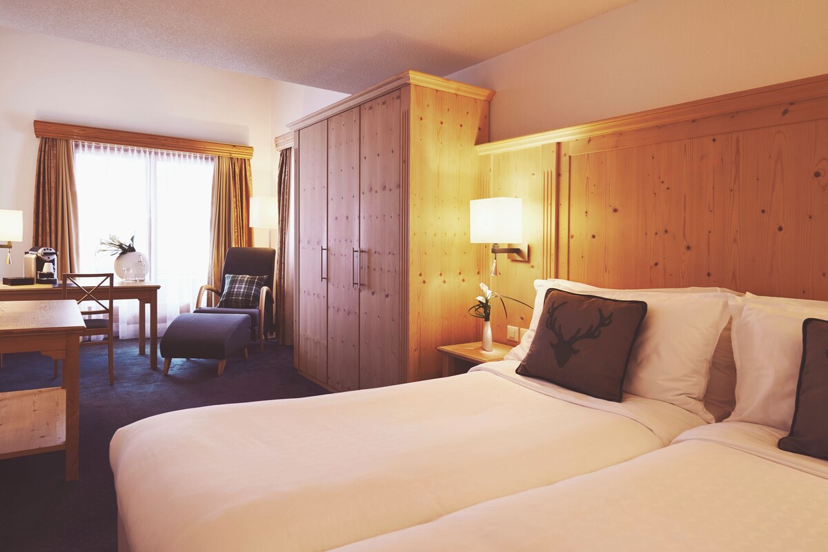 A hotel room with a large bed, a desk with a chair, a wardrobe, a bedside table with a lamp, and a window with curtains.